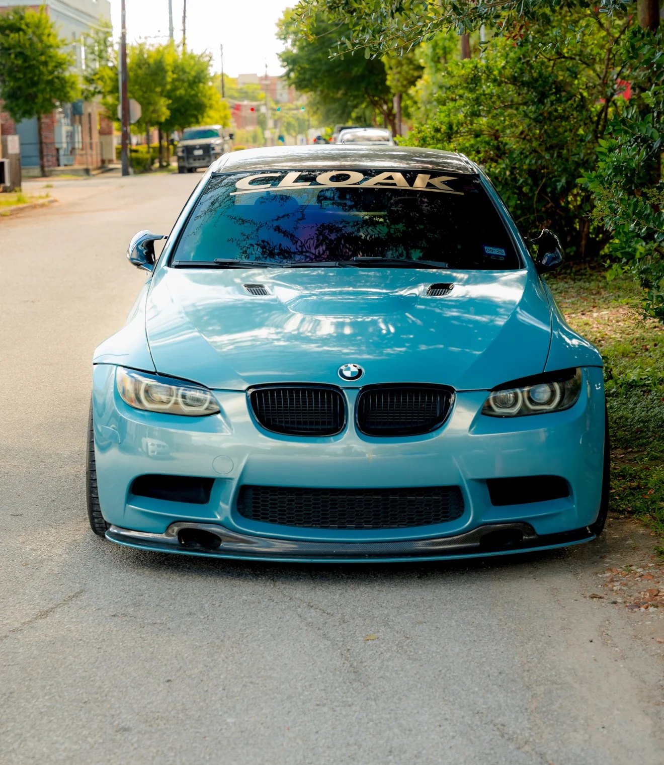 A light blue BMW M3 with the Cloak banner across its windshield, parked on a tree-lined street.