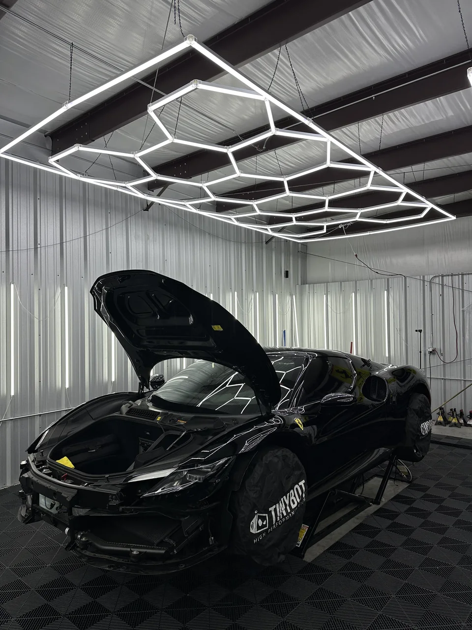 A black Ferrari 296 GTB in the Cloak showroom under the hexagon lights, hood up and front bumper off for PPF.