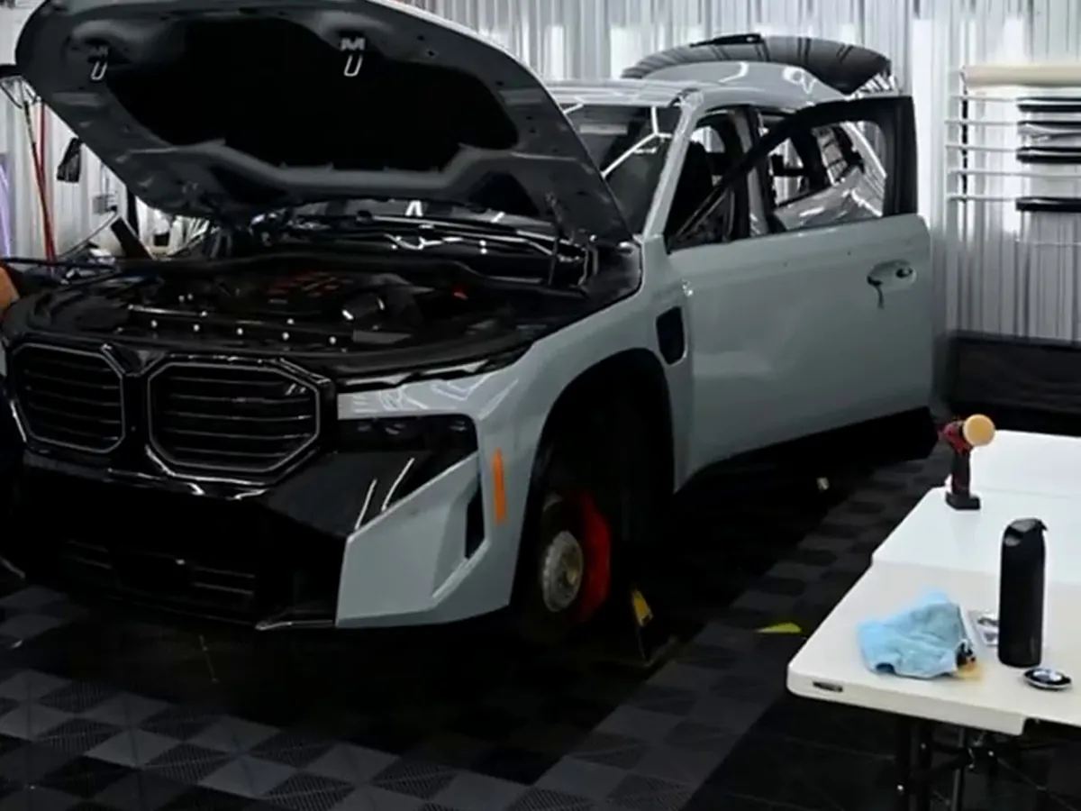 A light grey BMW XM in the showroom with its hood up and trim removed for wrapping.